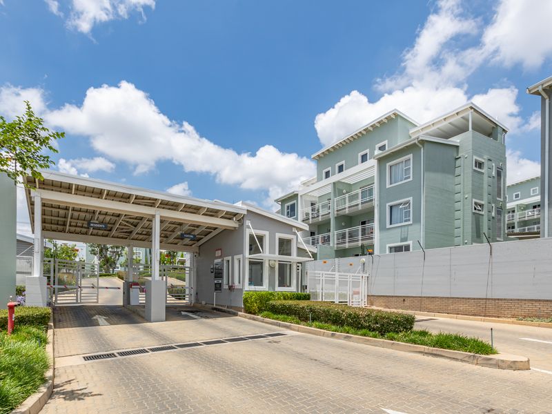 A well-maintained apartment complex entrance featuring a secure gatehouse, boom gate access, and paved driveway with perimeter electric fencing.