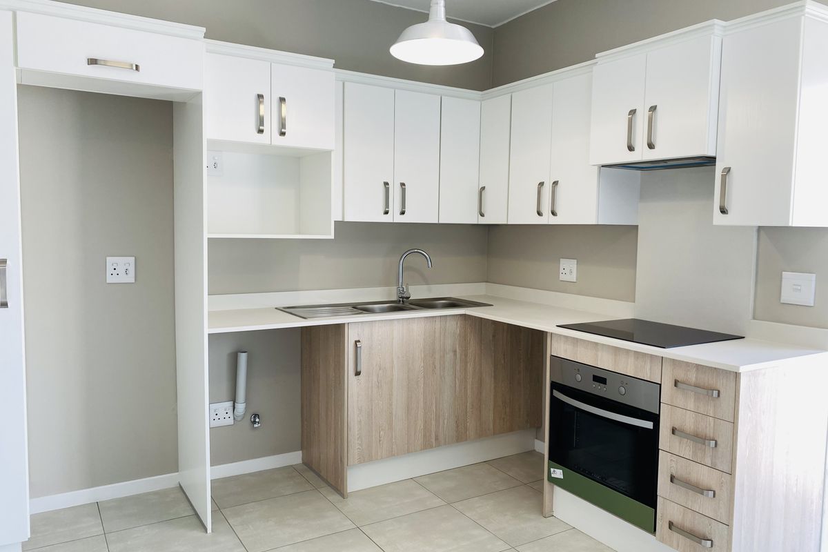 A modern, clean kitchen featuring white and wood-grain cabinetry, integrated electric oven and hob, stainless steel sink, and neutral tiled flooring.