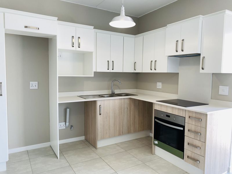 A modern, clean kitchen featuring white and wood-grain cabinetry, integrated electric oven and hob, stainless steel sink, and neutral tiled flooring.