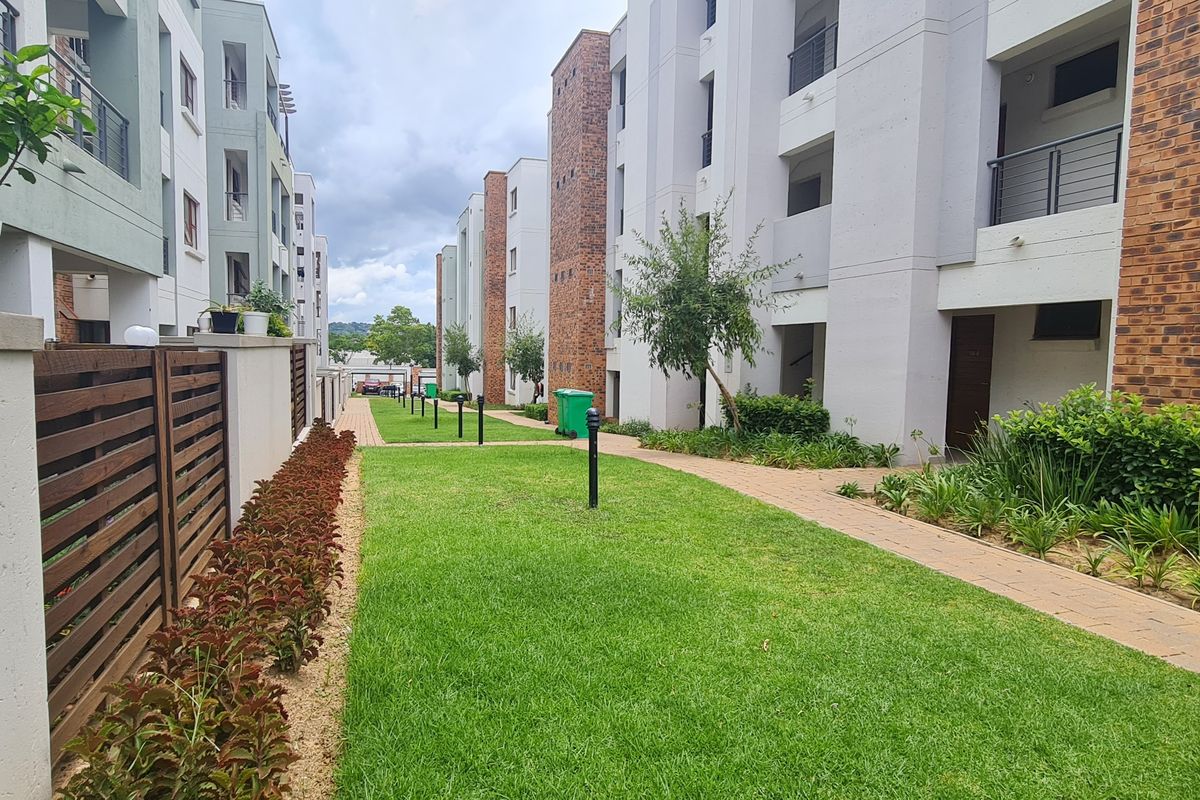 A well-maintained communal garden area between apartment blocks featuring paved walkways, manicured lawns, and modern exterior building finishes.