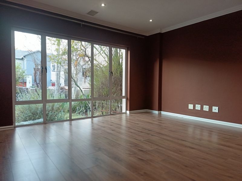 A spacious, empty living room featuring warm-toned laminate flooring, large windows with garden views, and recessed ceiling lighting.