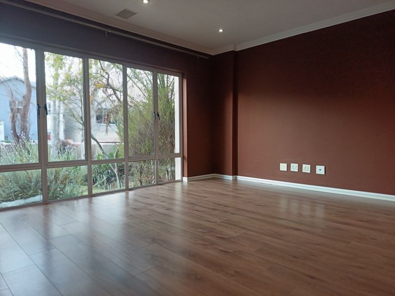 Spacious, empty living area featuring warm-toned laminate flooring, large windows providing natural light, and modern recessed downlighting.