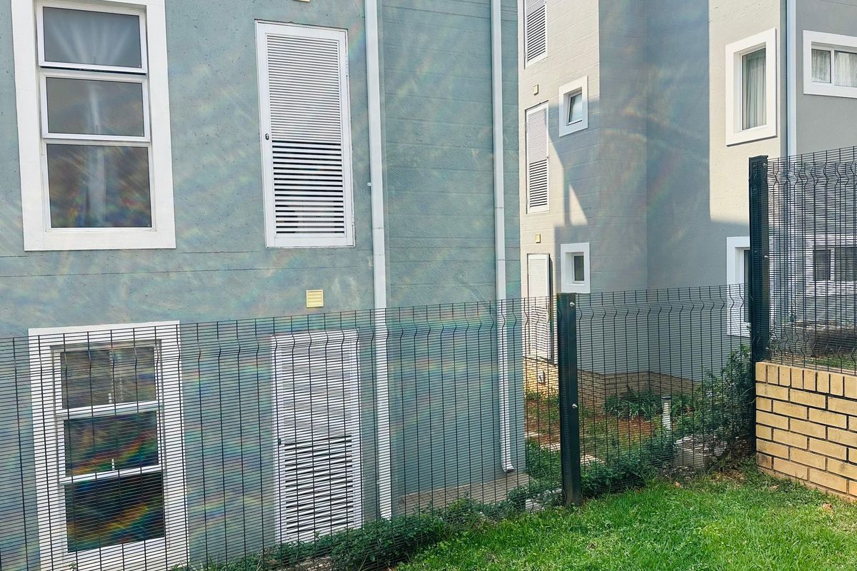 Rear exterior view of the apartment building featuring grey facade, louvered window shutters, and secure mesh perimeter fencing.