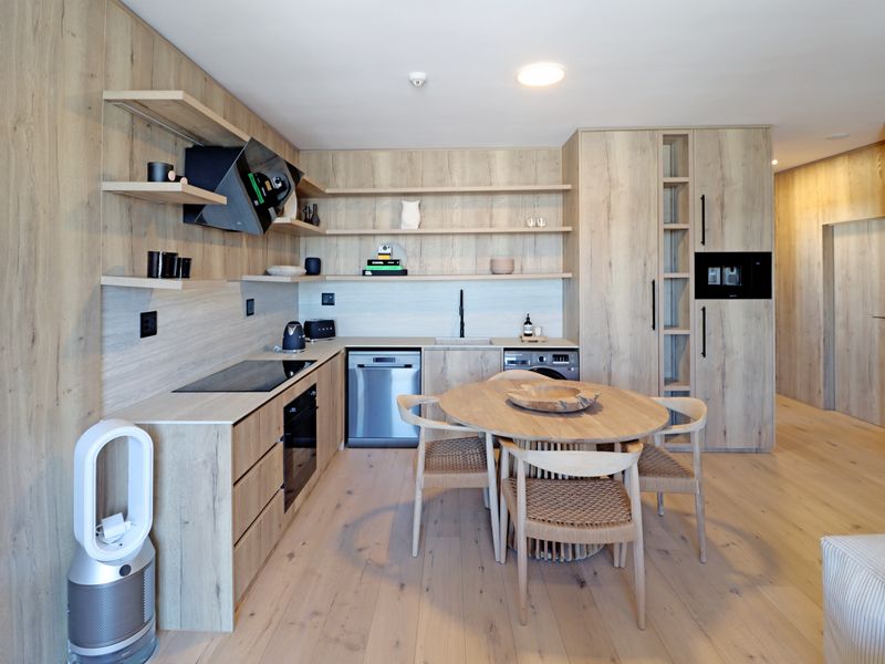 Modern open-plan kitchen featuring light wood cabinetry, integrated appliances, sleek shelving, and a dedicated dining area with timber flooring throughout.