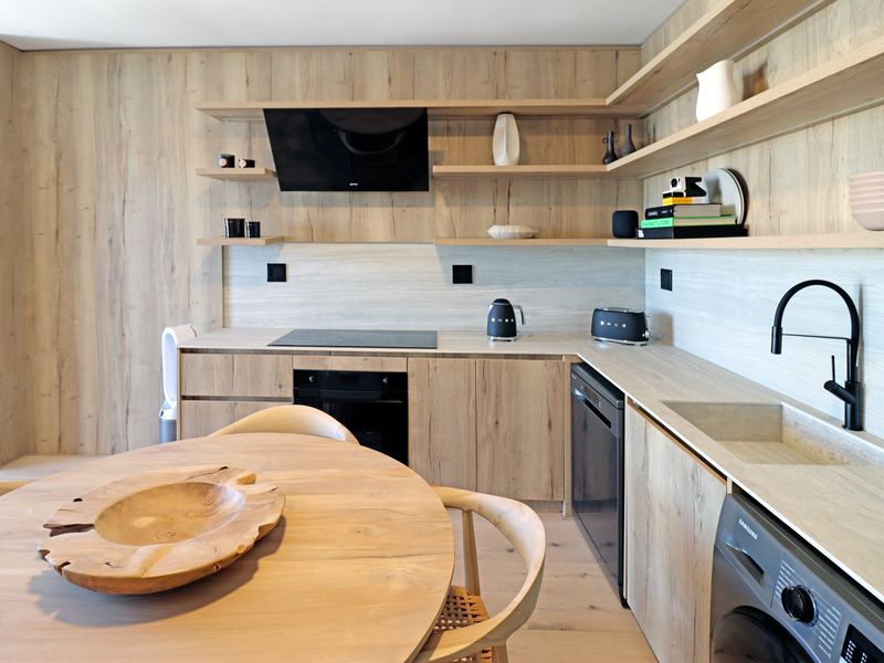 Modern open-plan kitchen featuring light wood cabinetry, integrated appliances, stone countertops, and a sleek black faucet, seamlessly blending with the dining area.