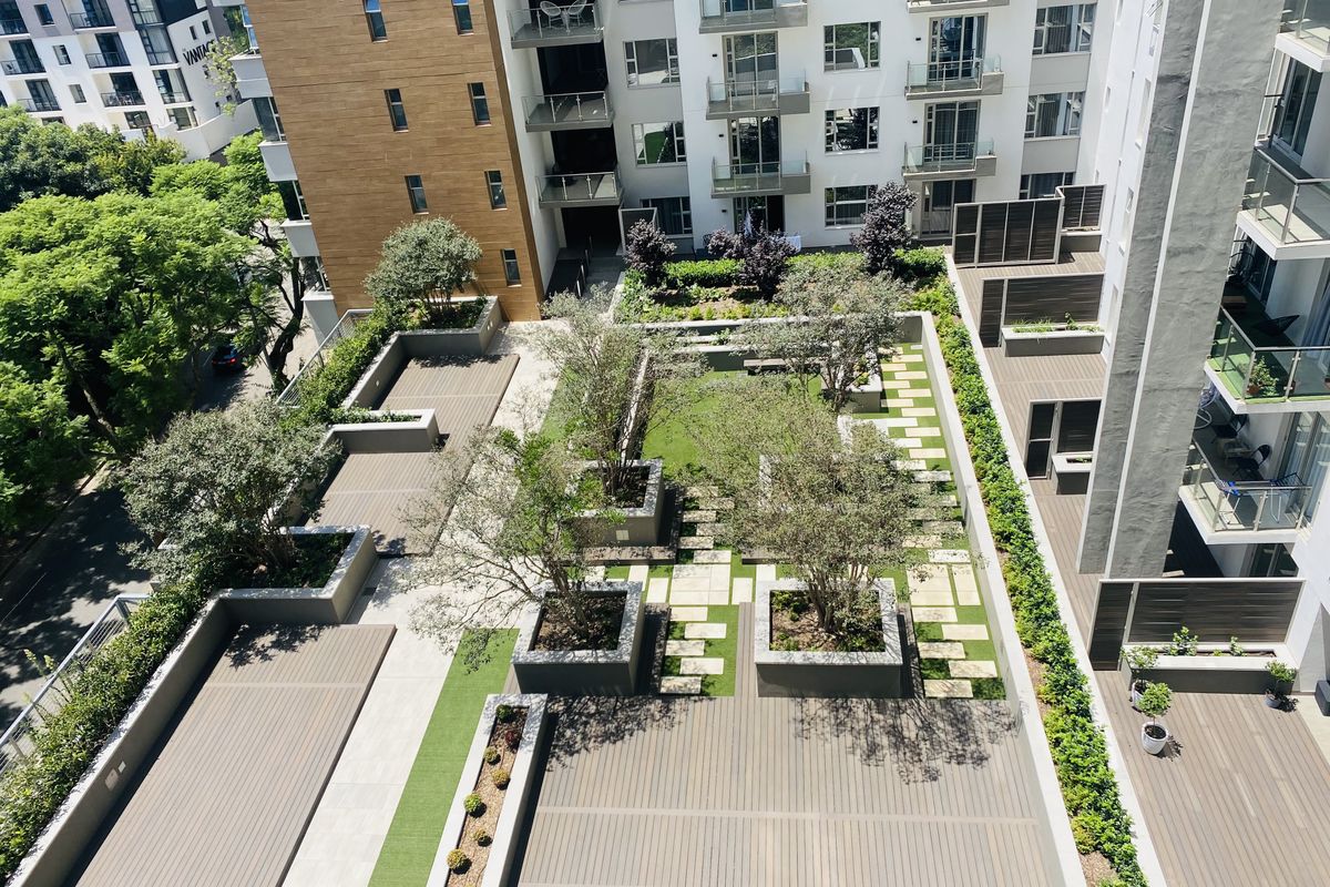 An elevated view of a modern apartment complex courtyard featuring landscaped gardens, paved walkways, raised planters, and timber decking.