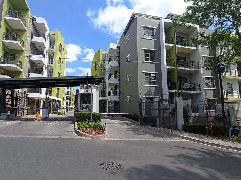 Exterior view of a modern apartment complex entrance featuring a secure boom gate, guard house, and multi-story residential buildings with balconies.