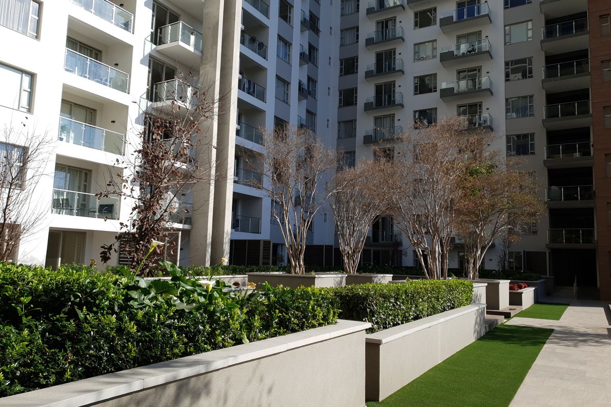 A well-maintained courtyard area between apartment blocks featuring raised garden beds, paved pathways, and artificial turf landscaping.