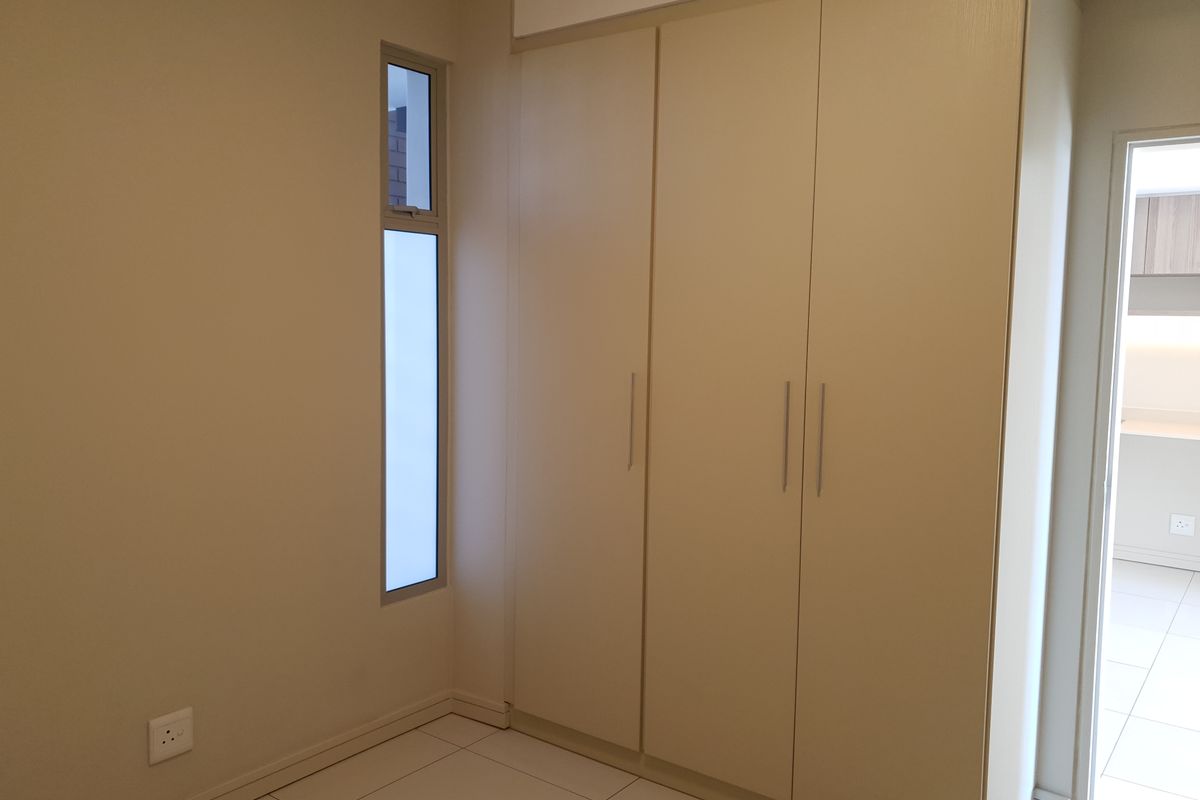 A clean, modern bedroom featuring neutral-toned built-in cupboards, tiled flooring, and a vertical window, offering a bright and minimalist living space.