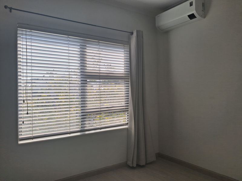 A bright, empty bedroom featuring laminate flooring, a wall-mounted split-system air conditioner, and window treatments including venetian blinds and a curtain rod.