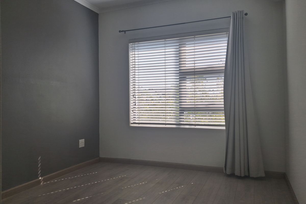 A bright, empty bedroom featuring modern laminate flooring, a grey accent wall, venetian blinds, and a wall-mounted air conditioning unit.
