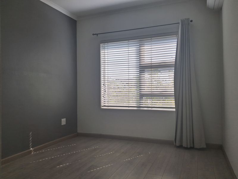 A bright, empty bedroom featuring modern laminate flooring, a grey accent wall, venetian blinds, and a wall-mounted air conditioning unit.
