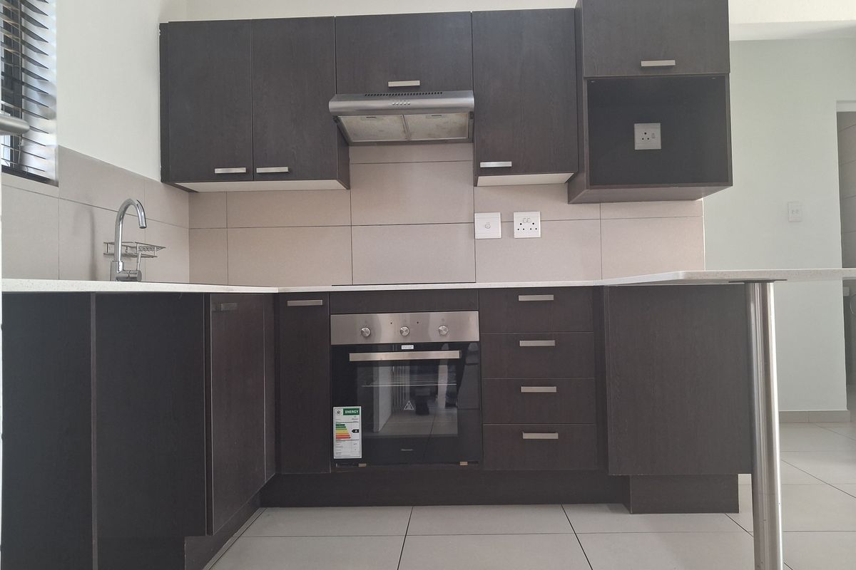 A modern, clean kitchen featuring dark wood cabinetry, stone countertops, a built-in oven, extractor fan, and neutral tiled flooring.