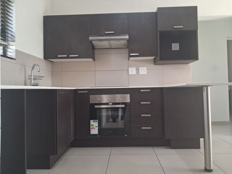 A modern, clean kitchen featuring dark wood cabinetry, stone countertops, a built-in oven, extractor fan, and neutral tiled flooring.