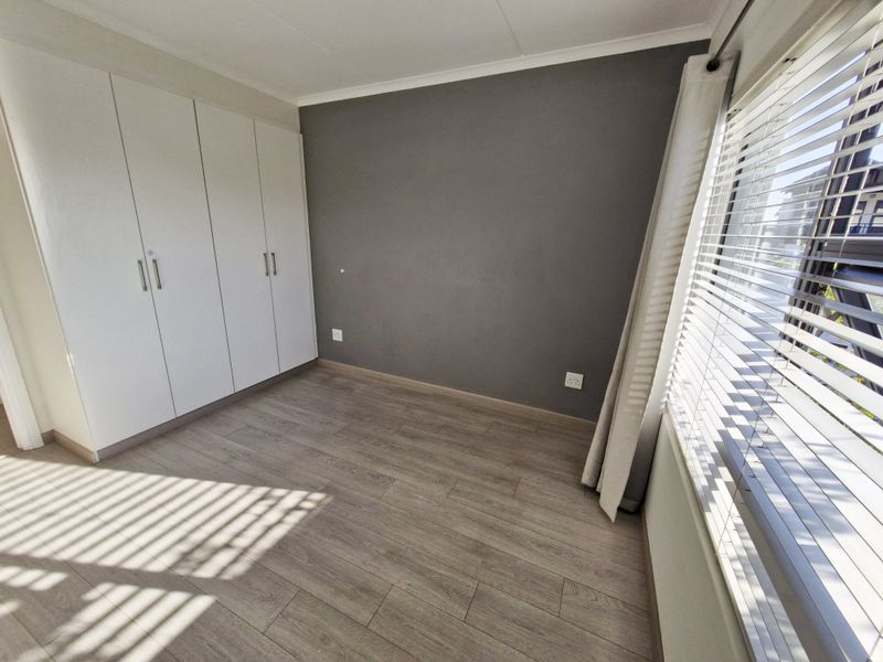 A bright, empty bedroom featuring modern laminate flooring, built-in white cupboards, and venetian blinds on the window.