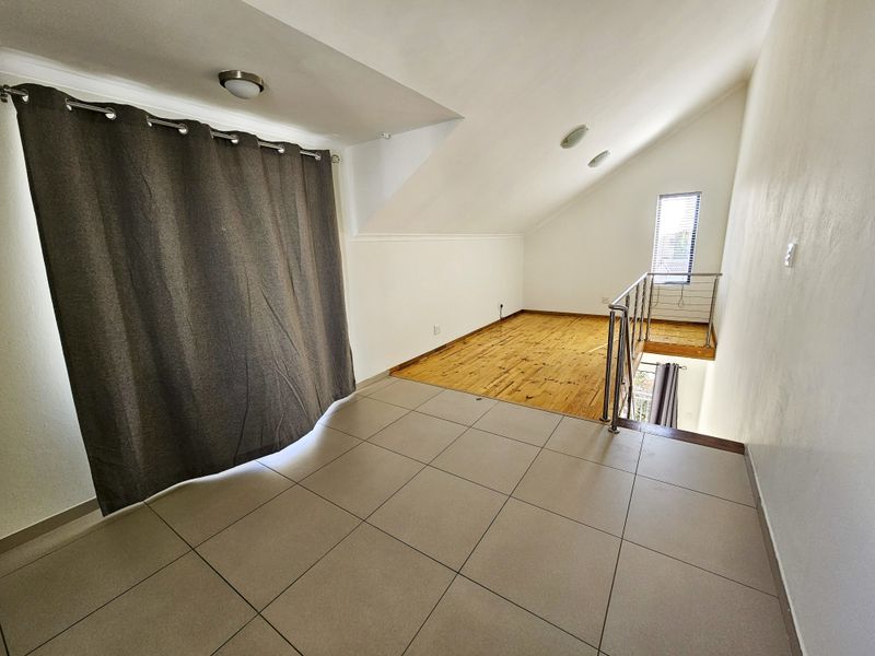A spacious loft-style bedroom featuring a mix of tiled and timber flooring, a stainless steel balustrade, and a sloped ceiling with recessed lighting.
