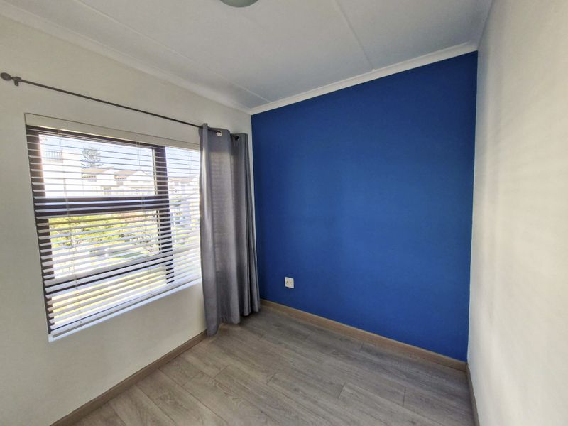 A bright, empty bedroom featuring modern laminate flooring, a large window with venetian blinds, and a bold blue accent wall.