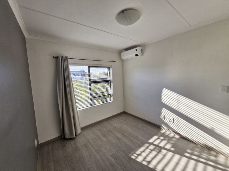A bright, empty bedroom featuring modern laminate flooring, an installed air conditioning unit, and large windows with venetian blinds.