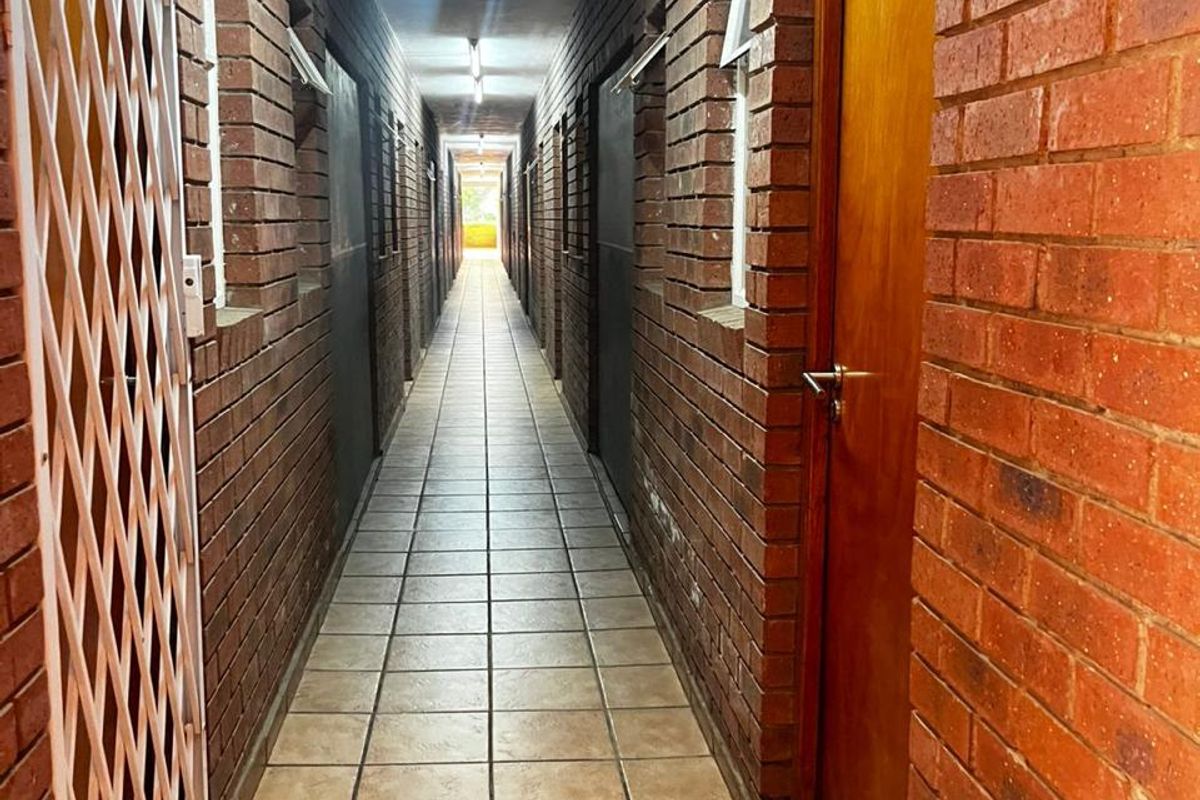 A long, narrow apartment hallway featuring exposed brick walls, ceramic tiled flooring, and multiple timber access doors with security gates.