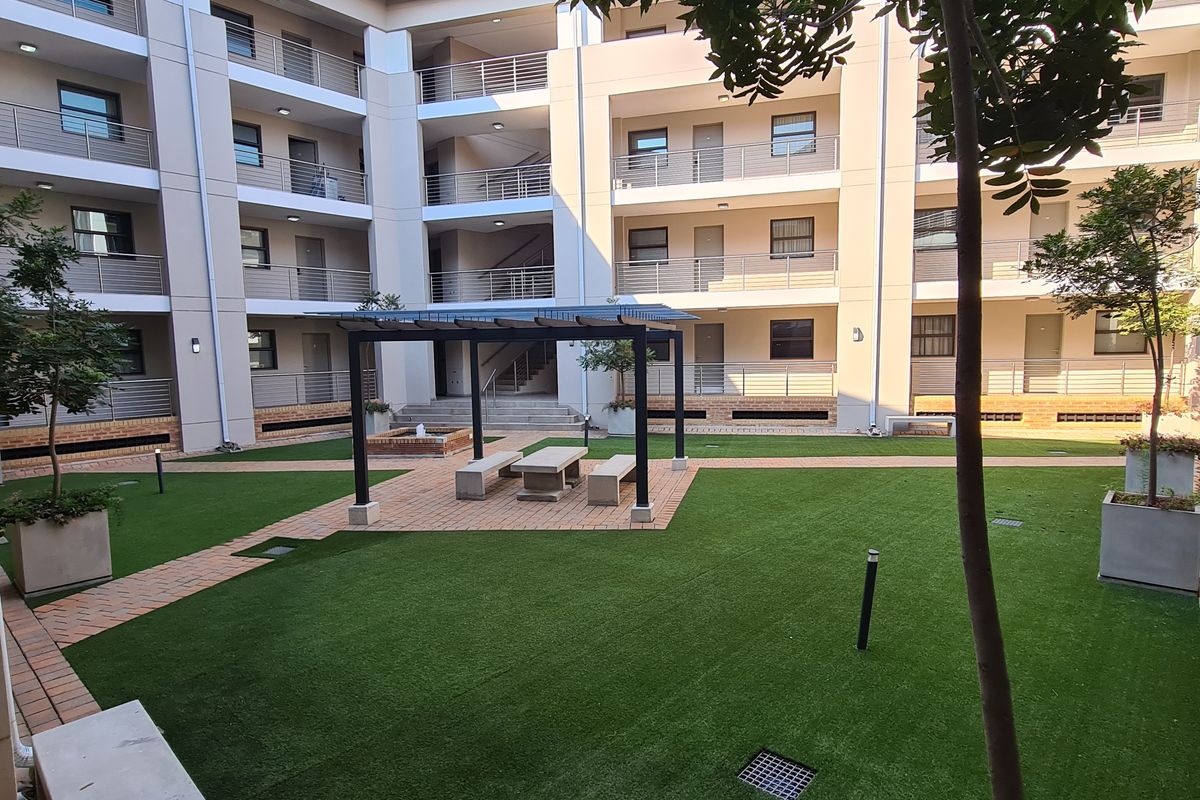 A well-maintained communal courtyard area within an apartment complex, featuring artificial turf, paved pathways, and a central pergola with built-in seating.