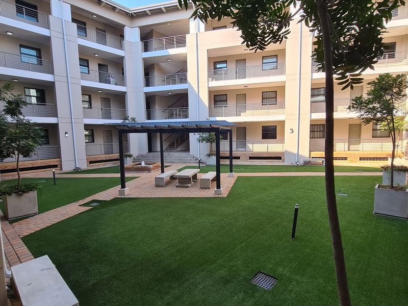 A well-maintained communal courtyard area within an apartment complex, featuring artificial turf, paved pathways, and a central pergola with built-in seating.