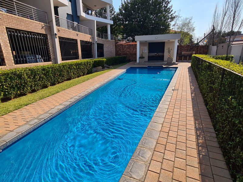 A well-maintained rectangular swimming pool area featuring paved decking, manicured hedges, and a poolside gazebo within a secure apartment complex.