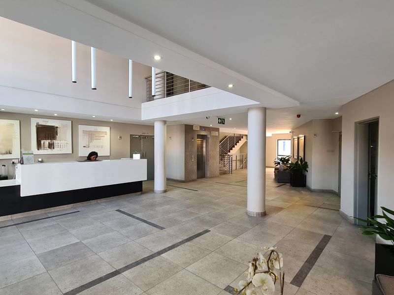 Spacious apartment building lobby featuring a reception desk, elevator access, tiled flooring, and a mezzanine level with a staircase.