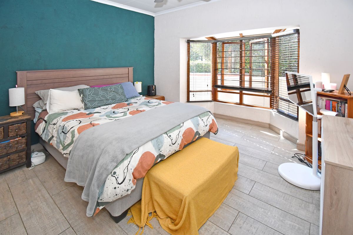 A well-lit bedroom featuring a large bay window with venetian blinds, tiled flooring, and a bold teal accent wall.
