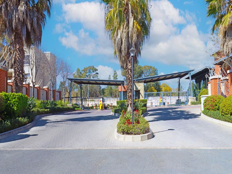 A well-maintained secure entrance to the apartment complex featuring a paved driveway, landscaped garden beds, and controlled security access gates.