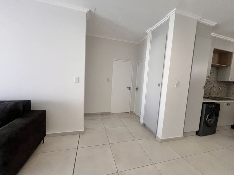 An open-plan living and kitchen area featuring neutral tiled flooring, clean white walls, and functional built-in cabinetry with integrated sink and laundry appliance space.