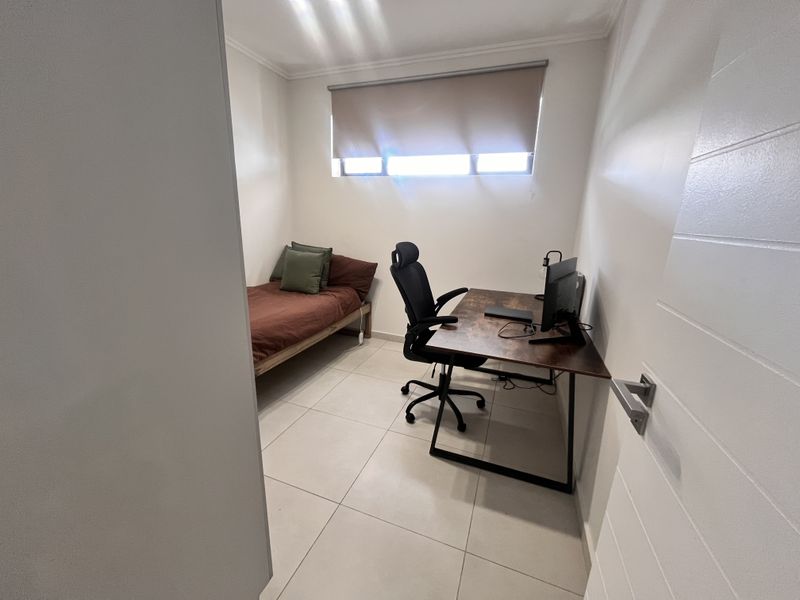 A compact bedroom featuring neutral tiled flooring and a roller blind, currently utilized as a combined sleeping and workspace area.