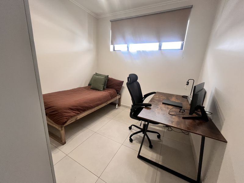 A compact bedroom featuring neutral walls, tiled flooring, and a roller blind, currently utilized as a combined sleeping and workspace area.