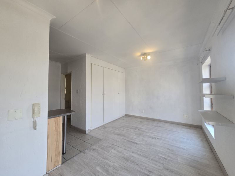 A bright, empty bedroom featuring light-toned laminate flooring, built-in cupboards, and floating wall shelves, offering a clean and versatile living space.