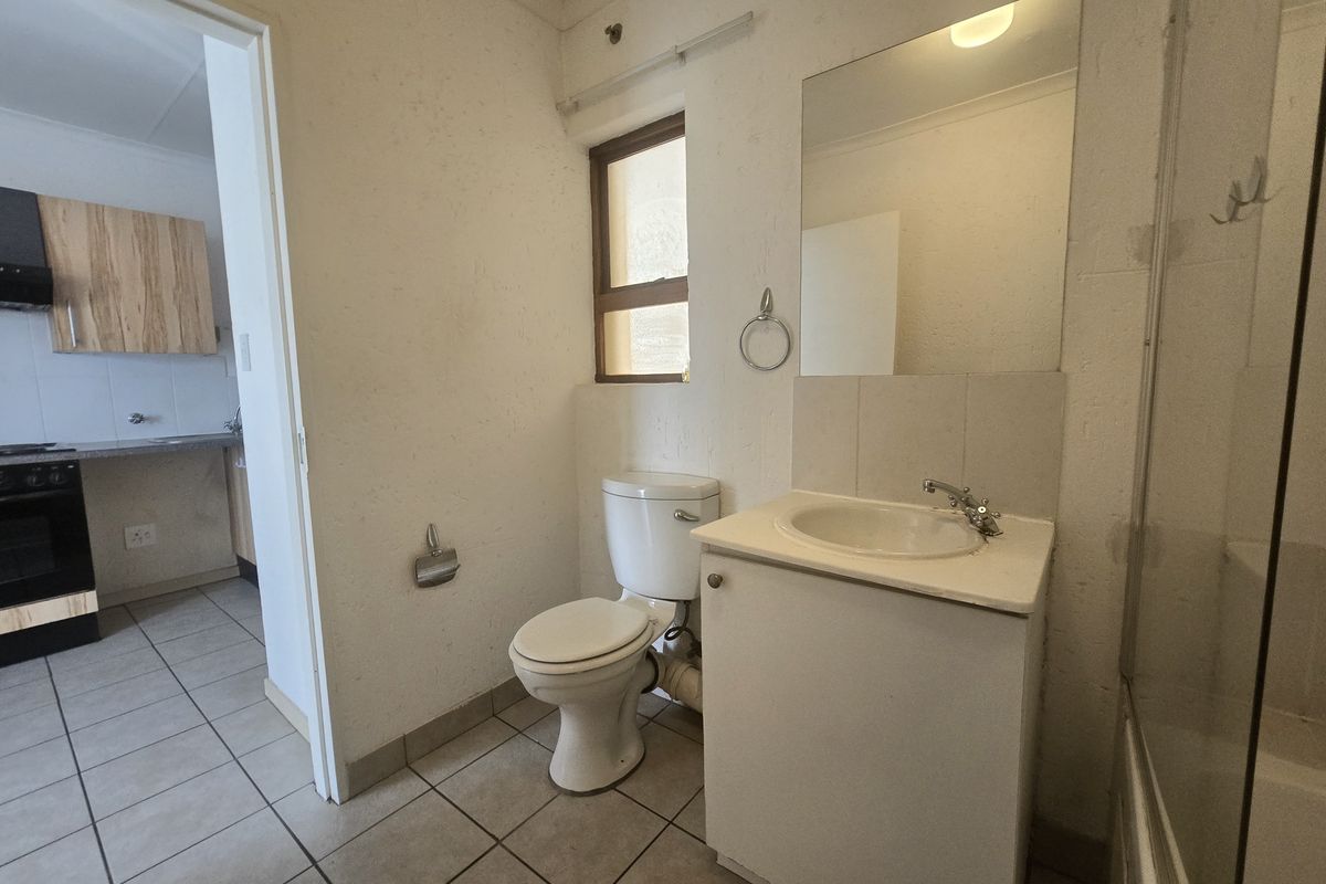 A functional bathroom featuring a vanity cabinet, basin, and toilet, with tiled flooring and a wall-mounted mirror, adjacent to the kitchen area.