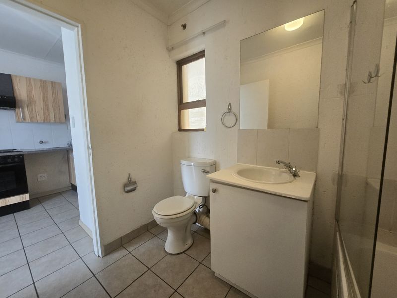 A functional bathroom featuring a vanity cabinet, basin, and toilet, with tiled flooring and a wall-mounted mirror, adjacent to the kitchen area.
