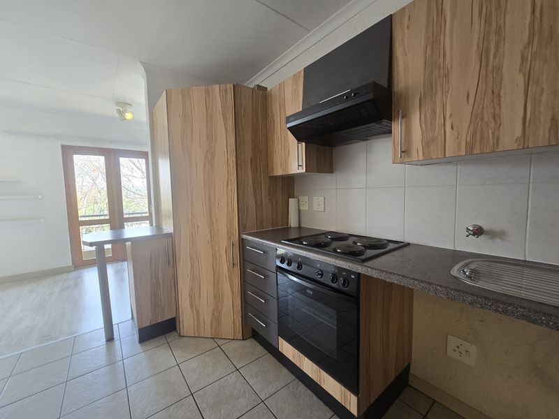 A functional kitchen featuring wood-look cabinetry, built-in oven and hob, tiled backsplash, and a breakfast bar, leading to an adjacent living area.
