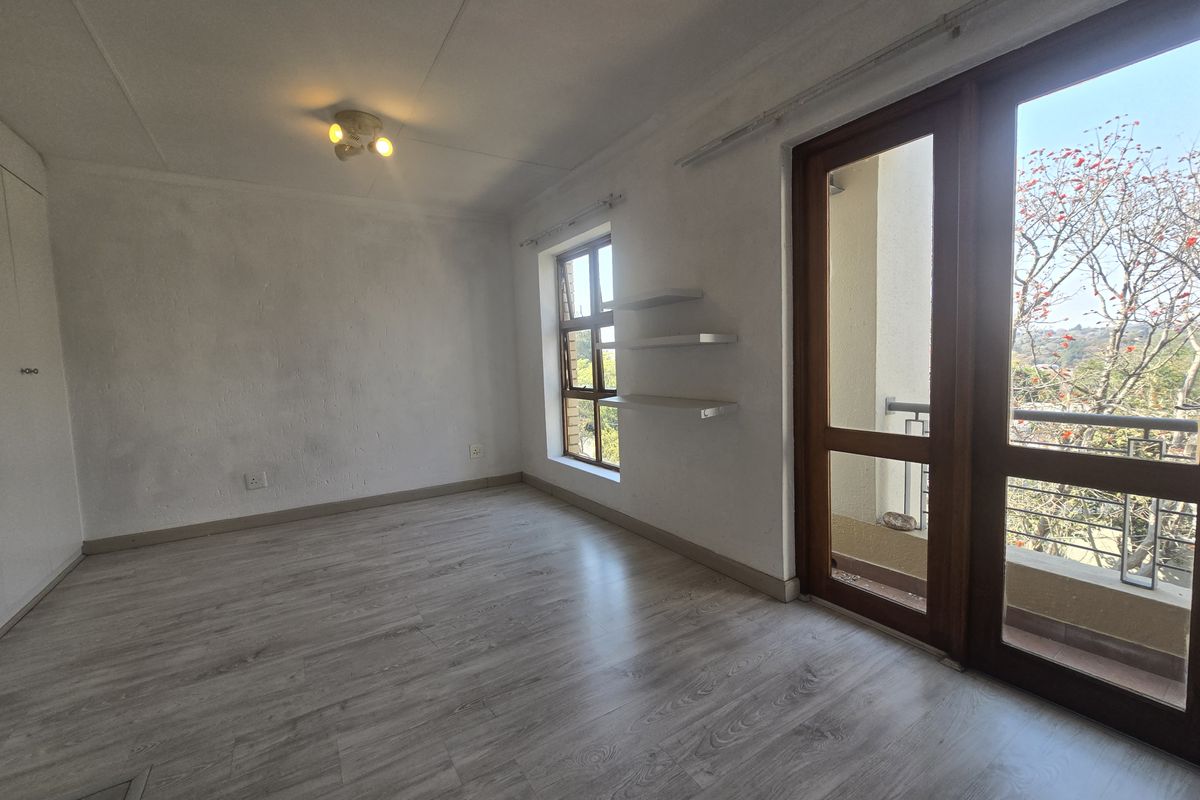 An empty bedroom featuring light-toned laminate flooring, built-in cupboards, floating shelves, and direct access to a balcony through a timber-framed glass door.