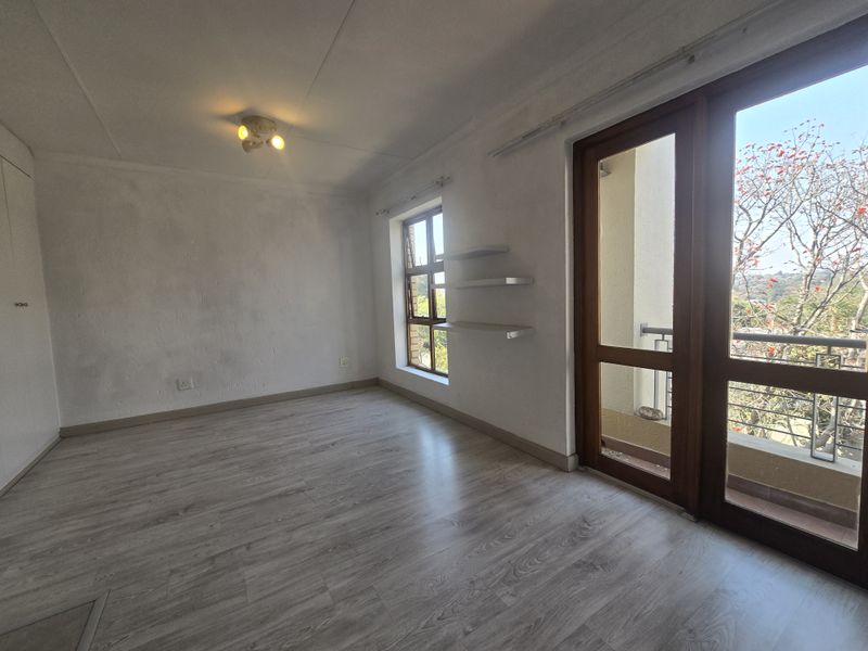 An empty bedroom featuring light-toned laminate flooring, built-in cupboards, floating shelves, and direct access to a balcony through a timber-framed glass door.