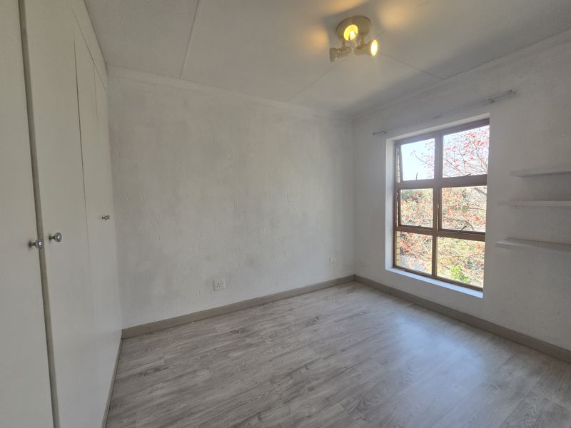 A bright, empty bedroom featuring modern laminate flooring, built-in cupboards, floating shelves, and a large window with natural light.
