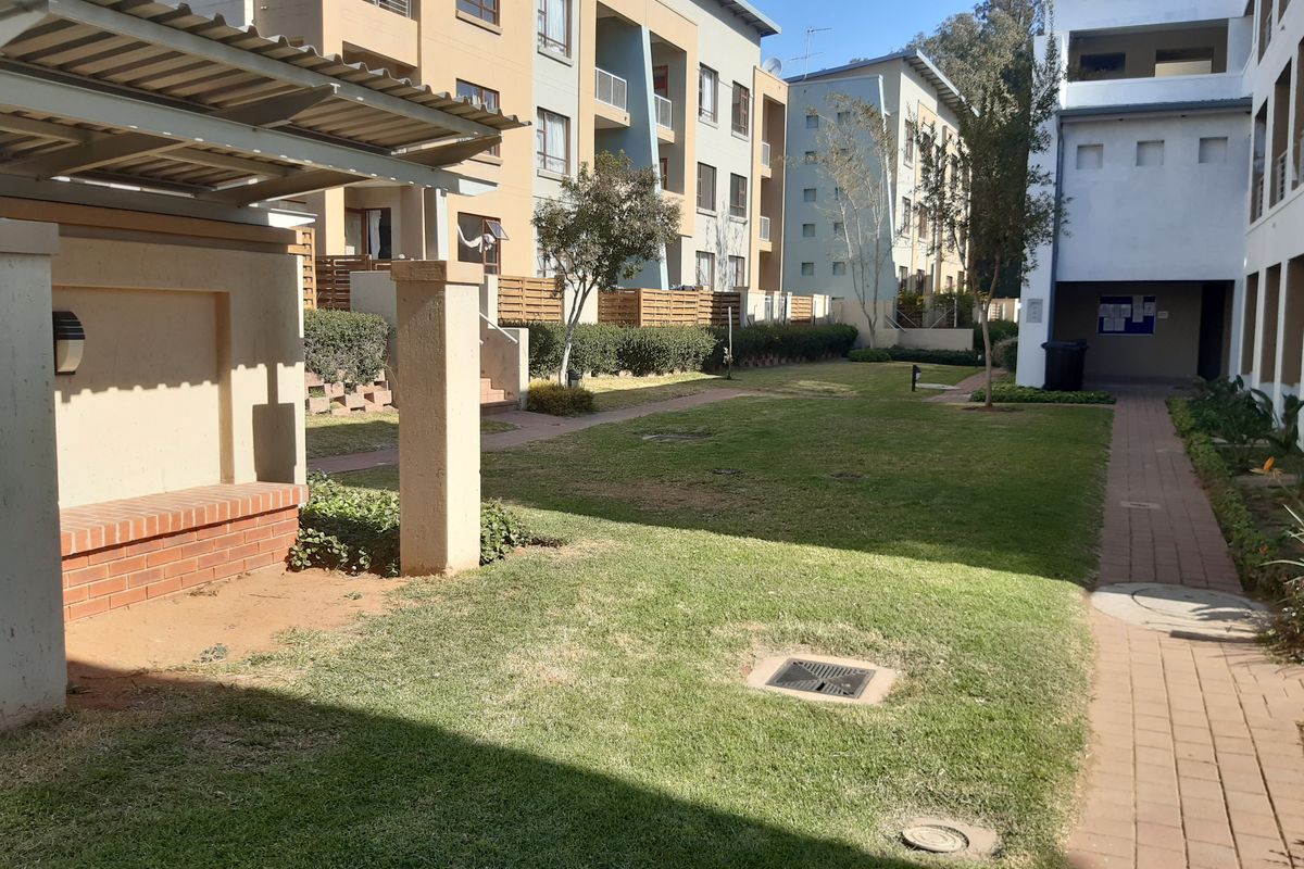 A well-maintained communal garden area featuring brick pathways, a covered seating structure, and landscaped lawn between apartment building blocks.