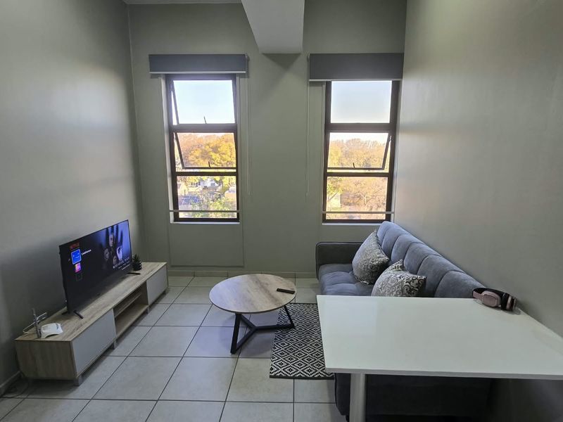 A well-lit living area featuring neutral wall tones, tiled flooring, and two large windows providing natural light and an exterior view.