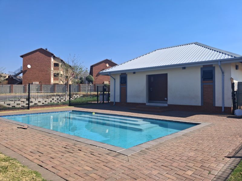 A well-maintained outdoor pool area featuring a clear swimming pool, brick paved surround, and a detached structure with a metal roof.
