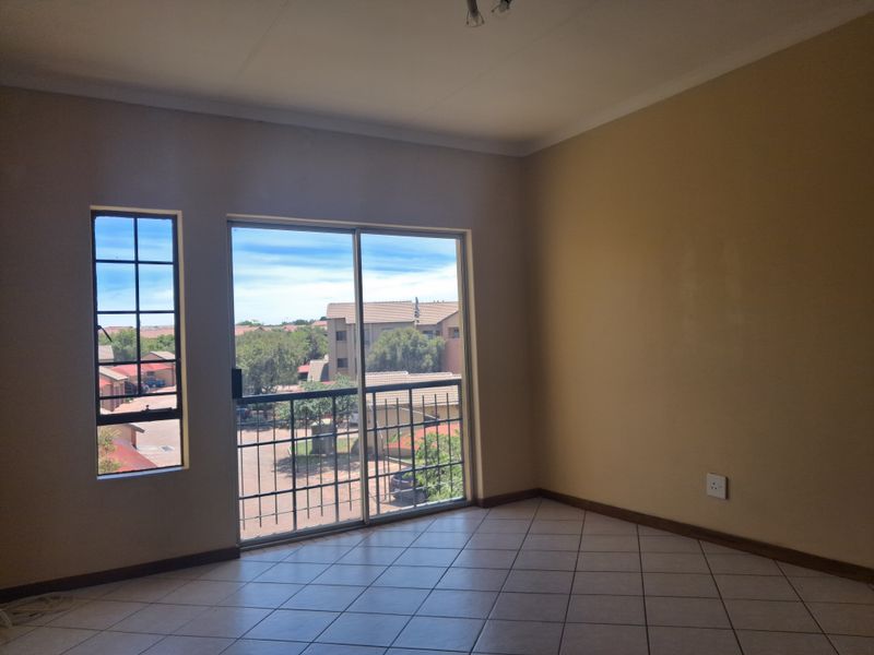 An empty living room featuring tiled flooring, a sliding glass door with security gate, and a casement window with views of the complex.