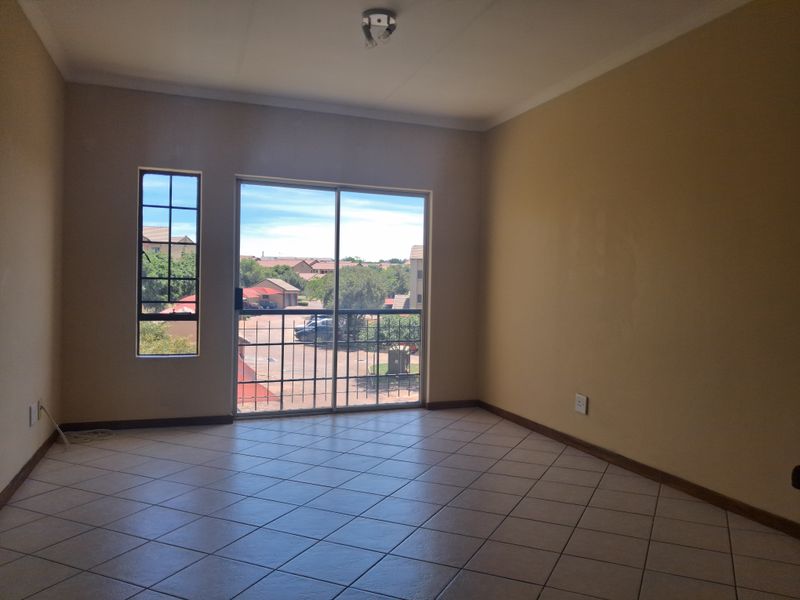 An empty, sunlit living room featuring neutral tiled flooring, a sliding glass door with security gate, and views of the surrounding townhouse complex.