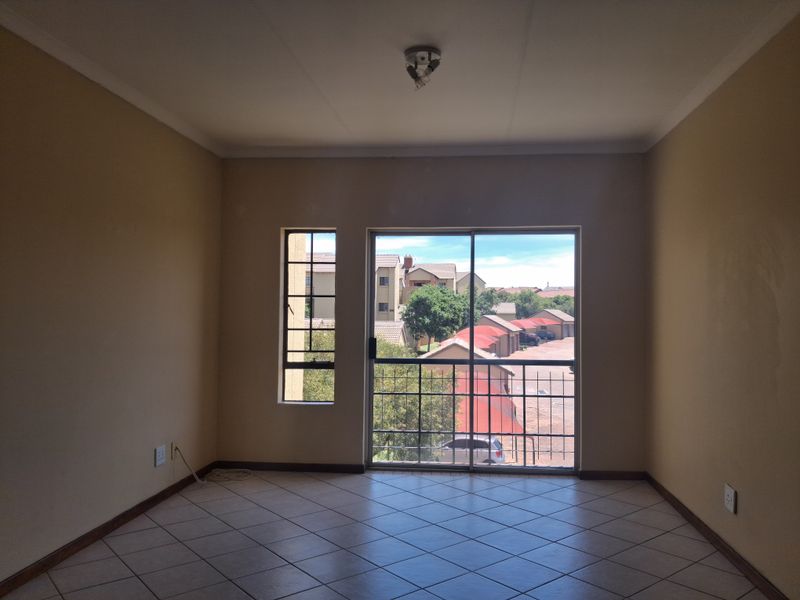 An empty living room featuring tiled flooring, a sliding glass door with security gate, and a window providing natural light and neighborhood views.