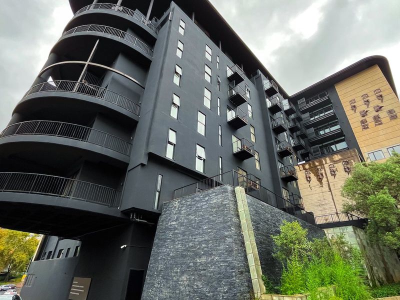 A modern multi-story apartment building featuring a dark facade, prominent curved balconies, and stone-clad retaining walls.