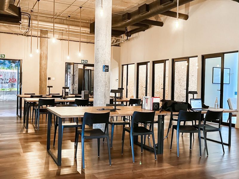 A modern open-plan office space featuring exposed industrial ceilings, concrete columns, timber flooring, and glass-partitioned meeting rooms.