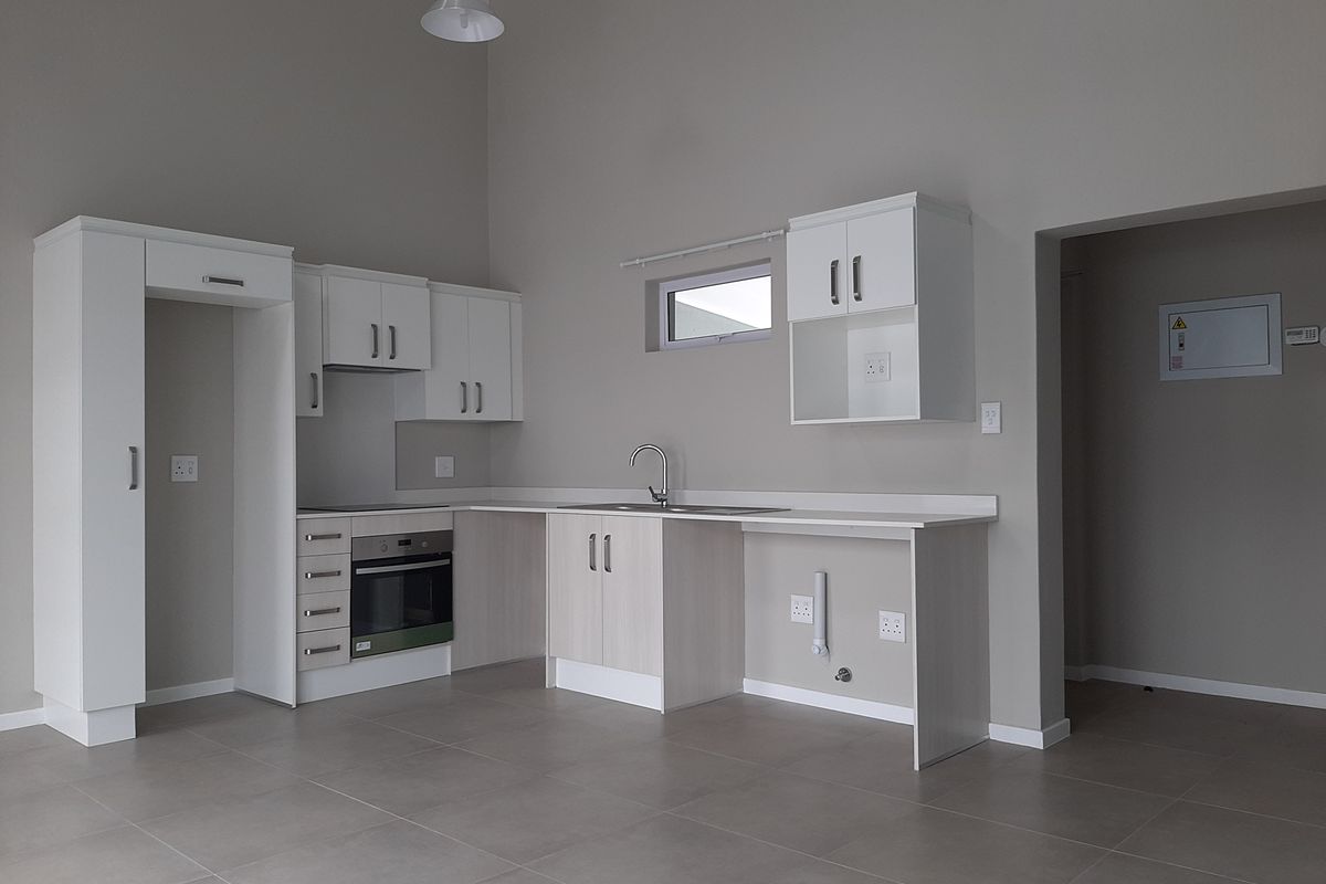 A modern, empty kitchen featuring white cabinetry, built-in oven and hob, stainless steel sink, and neutral tiled flooring in a clean, minimalist space.