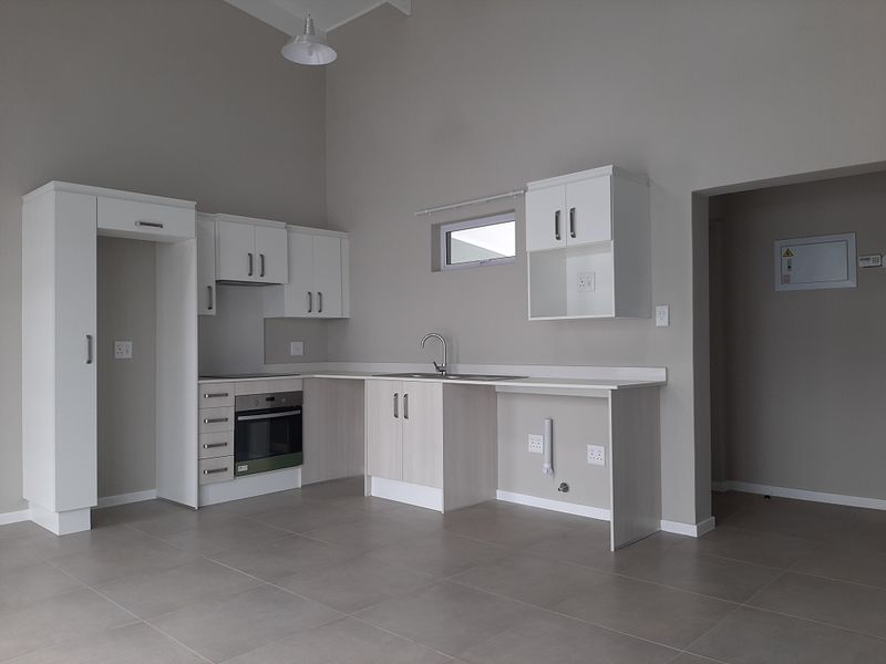 A modern, empty kitchen featuring white cabinetry, built-in oven and hob, stainless steel sink, and neutral tiled flooring in a clean, minimalist space.