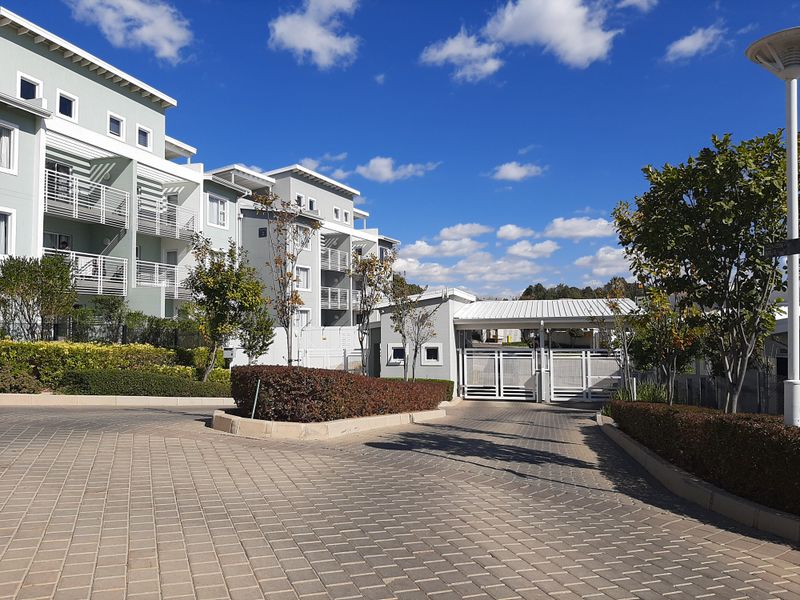 View of the apartment complex entrance featuring a paved driveway, security gate, guard house, and multi-story residential buildings with balconies.
